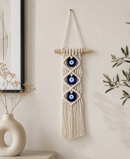 Cream macramè wall hanging with three navy blue evil eye discs on a wooden dowel styled on a white wall with dried flowers – THE AURA CO.