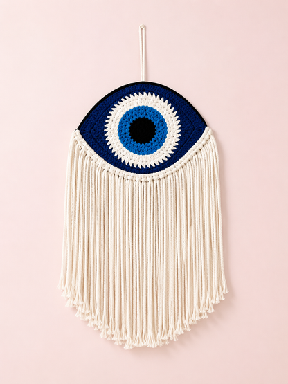 Cream macramè wall hanging with woven navy and blue evil eye design and long fringe on a blush pink background – THE AURA CO.